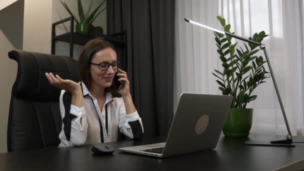 Gelukkig glimlachende vrouw spreekt per smartphone voor laptop zittend in modern kantoor. Zakenvrouw praat over de telefoon, met behulp van laptop zitten thuis bureau — Stockvideo