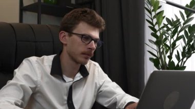 Gözlüklü genç işadamı ofiste dizüstü bilgisayar kullanıyor. Kendine güveni olan adam bilgisayarda çalışır, ekrana bakar ve modern ofis masasındaki klavyeye yazar.