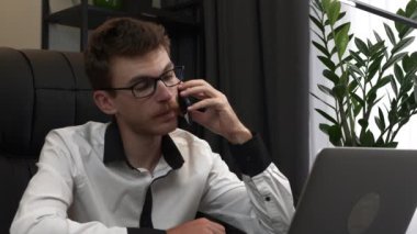 Neşeli genç iş adamı modern ofiste duygusal olarak cep telefonuyla konuşuyor. Mutlu adam ofis masasında akıllı telefonla konuşuyor. Gülümseyen erkek iş yerinde cep telefonu kullanıyor