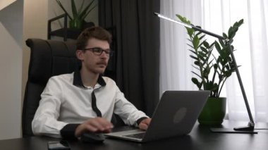 Gözlüklü akıllı adam modern ofiste dizüstü bilgisayar kullanıyor. İş adamı ev işyerindeki bilgisayarda çalışıyor. Çalışan dizüstü bilgisayarda çalışır, ekrana bakar ve ofis masasındaki klavyeye yazar.