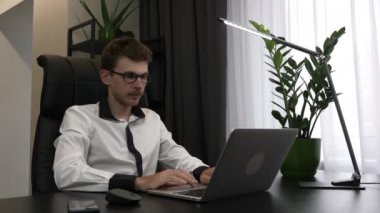 Gözlüklü ve gömlekli genç adam iş yerinde oturuyor, dizüstü bilgisayarı açıyor ve çalışmaya başlıyor. İşadamı modern ofiste bilgisayar kullanıyor. Erkek bilgisayarda çalışır.