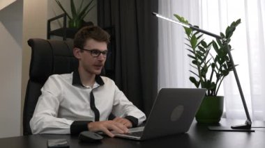 İş adamı, iş yerindeki çalışma ekibinin çevrimiçi video konferansını yapıyor. Genç erkek serbest çalışan web kamerası ile konuşuyor, dizüstü bilgisayara bakıyor ve gülümsüyor. Adam video aracılığıyla konuşuyor.