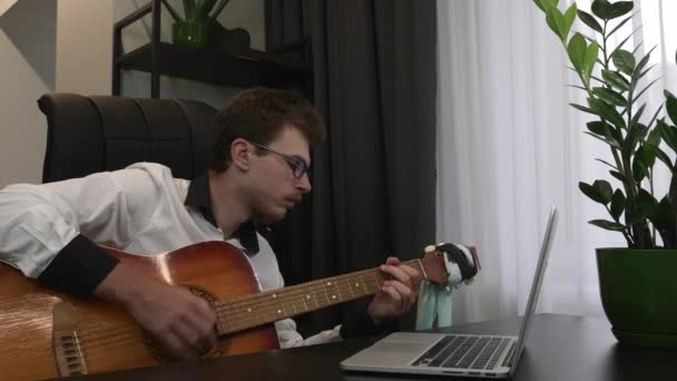 Уверенный в себе молодой тренер по игре на гитаре смотрит на экран ноутбука и записывает онлайн уроки музыки через веб-камеру. Кавказский музыкант играет на акустической гитаре в офисе. — стоковое видео