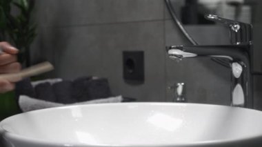 Banyodaki kadın diş fırçasına diş macunu sürüyor, dişlerini fırçalamaya hazırlanıyor. Kadın diş macununu fırçaya koyar, lavabonun üzerinde tutar.