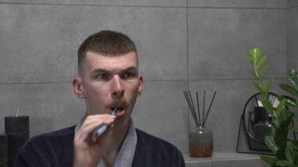 早上，穿着浴衣的快乐年轻人正在现代浴室里用牙刷和牙膏刷牙。英俊的男性刷牙，早上做口腔保健常规 — 图库视频影像
