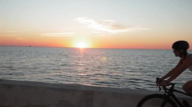 Kadın gün batımında arka planda denizle bisiklet sürüyor. Günlük kıyafetleri ve şık gözlükleri olan bayan bisikletçi gün doğumunda deniz kenarında bisiklet sürüyor.