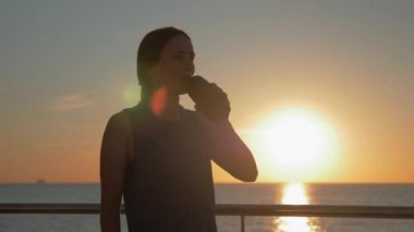Genç, çekici, gülümseyen bayan atlet antrenmandan önce gün doğumunda su içiyor. Esmer kadın gün batımında sahilde ayakta su içiyor. Koşmak ve sağlıklı yaşam tarzı kavramı