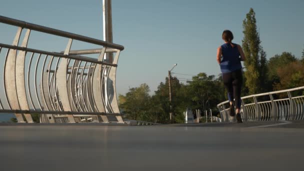 Fitness žena s krásným sportovním tělem je běh podél pláže promenády při západu slunce. Motivovaný ženský běžec běhá ráno na mostě a provádí kardiochirurgická cvičení jako součást každodenní rutiny — Stock video