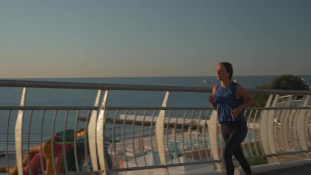 Glad attraktiv ung kvinna i blå sportkläder körs på bron på morgonen vid soluppgången. Leende brunett kvinnliga idrottare löper längs strandpromenaden i solljus. Friska livsstil koncept — Stockvideo