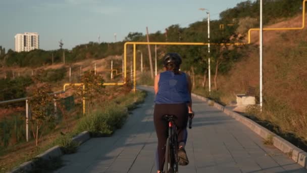 Mujer en ropa deportiva azul paseos en bicicleta a lo largo de carril bici en el soleado día de verano. Mujer ciclista en casco y ropa de ciclismo está pedaleando en bicicleta de carretera por la mañana. Concepto de deporte y ciclismo — Vídeos de Stock