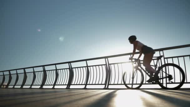 Велосипедистка в стильном наряде и шлеме крутит педали на дорожном аэробайке в солнечных лучах. Женщина-спортсмен крутит педали на велосипеде по велосипедному мосту на закате. Счастливая девушка ездит на велосипеде — стоковое видео