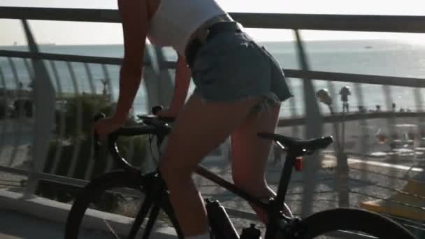 Vrouw in top en jeans shorts rijdt op de fiets langs de strandpromenade, van dichtbij. Hipster vrouwelijke fietser fietst op de racefiets bij zonsondergang. Happy lifestyle concept — Stockvideo