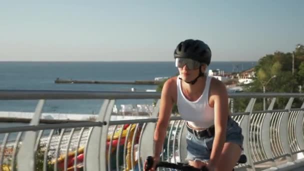 Joven morena en forma en casco profesional y gafas está pedaleando en bicicleta en el paseo marítimo al amanecer bajo la luz del sol. Elegante chica sonriente feliz está disfrutando de paseo en bicicleta a lo largo de la orilla del mar — Vídeos de Stock