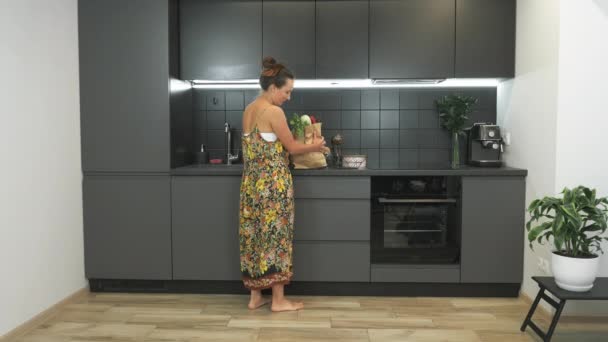 Giovane bella donna sta disimballando verdure e prodotti dal negozio in cucina moderna a casa. Bruna affascinante femmina è in cucina con pacchetto di prodotti freschi in mano — Video Stock