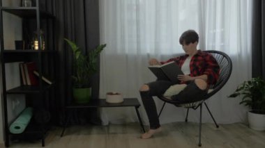 Gündelik giysiler giyen Milenyum tarzı bir kız modern oturma odasında rahat bir sandalyeye oturmuş kitap okuyor ve iş gününden sonra dinleniyor. Esmer kadın kitap okuyor, hafta sonunun keyfini çıkarıyor.