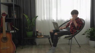 Heyecanlı genç kadın dizüstü bilgisayar ekranına bakıyor, klavyede yazıyor ve evdeki koltukta çay içiyor. Kız öğrenci e-postayla haber okuyor, online eğitim ve dersler izliyor, internetten okuyor.