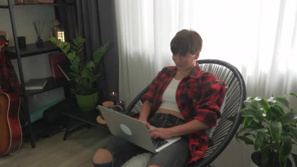 Frau sitzt auf gemütlichem Stuhl in ihrer modernen Wohnung, tippt Nachrichten auf Laptop-Tastatur, chattet online in sozialen Medien mit Freunden. Frau benutzt Notizbuch von zu Hause aus und macht Remote-Projekt — Stockvideo