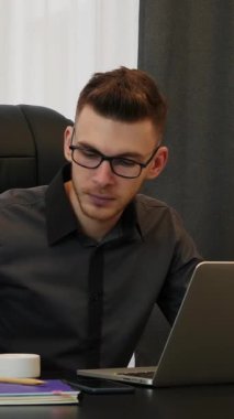 Modern ofisinde kahve içen bir adam. Takım elbiseli ve bardaklı bir işadamı iş yerinde kahve içer. Erkek ofis masasında oturup sıcak bir şeyler yudumluyor. Dikey video