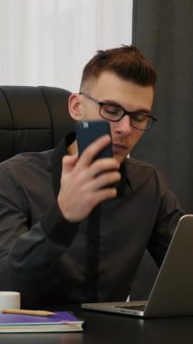 Telefondaki adam dizüstü bilgisayarın önünde oturuyor. Modern iş yerinde gözlüklü bir erkek akıllı telefondan konuşur. Ofiste oturan ve ortaklarıyla telefonla konuşan biri. Dikey video