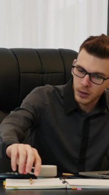 Gözlüklü bir adam ofiste bilgisayarın önünde oturmuş defterine not alıyor ve şirketin mali raporlarına bakıyor. Dikey video