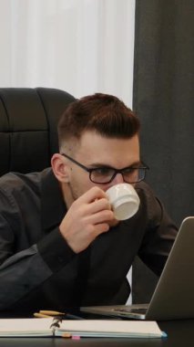 Modern ofiste işyerinde oturan adam kahve espresso içiyor. Takım elbiseli ve bardaklı bir erkek dizüstü bilgisayarın önünde kahve içiyor. Dikey video