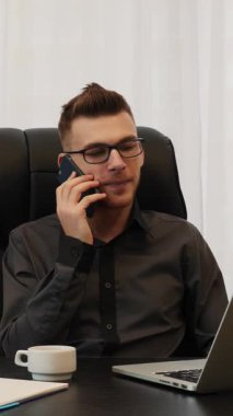 Adam telefonda ortaklarıyla konuşuyor, dizüstü bilgisayarın önünde oturuyor. Ofiste oturan erkek işçi, bilgisayar ekranına bakıyor ve akıllı telefondan konuşuyor. Dikey