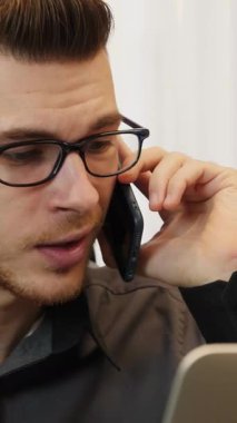 Modern ofiste sinirli bir adam telefonda konuşuyor. Sinirli erkek dizüstü bilgisayarın önünde oturuyor ve akıllı telefonla konuşuyor. İş yerinde sandalyede oturuyor. Dikey