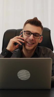 Modern ofiste dizüstü bilgisayarın önünde oturan bir işçi, iş arkadaşlarıyla akıllı telefondan konuşuyor. İşteyken birden fazla iş. Dikey video