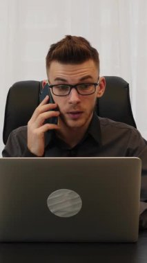 Dizüstü bilgisayarın önünde oturan ve akıllı telefondan konuşan bir adam. Gözlüklü ve gömlekli bir erkek ofiste oturuyor, dizüstü bilgisayara bakıyor ve telefonda konuşuyor. Dikey video