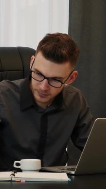 Ofiste çalışan bir adam, dizüstü bilgisayara bakıyor, not alıyor ve gülümsüyor. Güvenilir odaklı adam mali rapor tutar. Dikey video