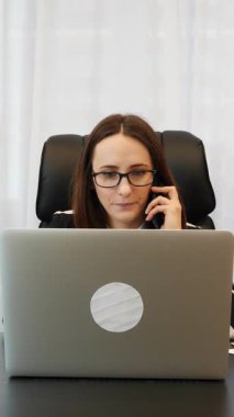 Modern ofisteki çalışma masasında oturan ve akıllı telefondan konuşan bir kadın. Dizüstü bilgisayarda çalışan ve telefonda konuşan bir kadın. Dikey video