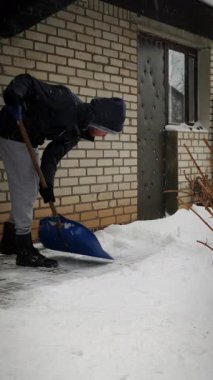 Adam kar fırtınasında arka bahçede kar kürüyor. Erkek kışın evin yanındaki karları temizler. Dikey video