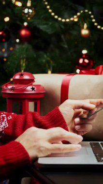 Kırmızı Noel kazaklı kadın kredi kartı tutuyor ve yeni yıl için online alışveriş yapıyor. Kadın Noel ağacının yanında oturuyor ve internetten hediyeler alıyor. Dikey video