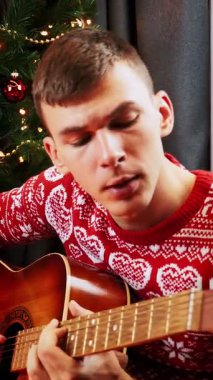 Kırmızı şenlikli süveter giyen, gitar çalan ve Noel şarkıları söyleyen gülümseyen bir adam. Yeni yıl ağacının yanında oturuyor. Yeni yıl 2025. Dikey video
