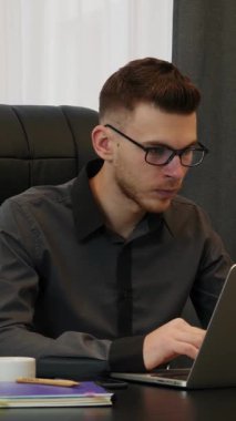 Ofiste oturan ve dizüstü bilgisayarda yazan gözlüklü bir adam. Modern ofiste bilgisayar üzerinde çalışan kendine güvenen genç bir erkek yönetici. Dikey video