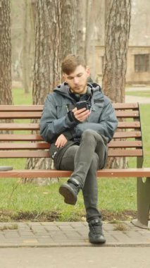 Sonbahar parkında oturan ve cep telefonunu sosyal ağ için kullanan bir adam. Dikey video