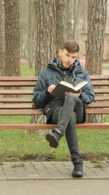 Genç adam kitap okuyor. Sonbahar parkında bankta oturuyor. Üzerinde mavi ceket var. Şehir parkında oturan, kitap okuyan ve sayfaları çeviren bir erkek. Dikey video