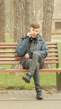 Sonbahar parkında bankta otururken telefonda konuşan yakışıklı bir adam. Dikey video