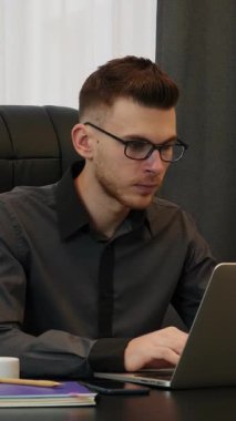 Ofiste dizüstü bilgisayarda çalışan bir adam. İş yerinde çalışan kendine güvenen genç bir adam. Tişört giyen ve gözlüklü bir erkek bilgisayarda yazı yazıyor. Dikey video