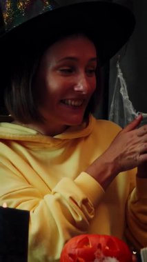 Cadılar Bayramı için dekore edilmiş odada elinde balkabağı tutan cadı şapkalı gülümseyen bir kadın. Jack-O-Lantern. Ürkütücü yüzlü balkabağı oyulmuş bir kadın. Dikey video