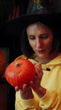 Cadılar Bayramı için turuncu balkabağı boyayan cadı şapkalı tatlı genç kız. Kadın Cadılar Bayramı partisi için hazırlanıyor. Mutlu cadılar bayramı, dekorasyon ve geleneksel semboller. Dikey video