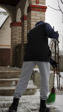 Sıcak kış giysileri içindeki adam kışın evin yakınındaki karları süpürür. Genç erkek, kar fırtınasından yolu temizliyor. Dikey video