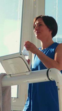 Sportif, genç, sağlıklı bir kadın güneşli bir günde pencereye karşı koşu bandında egzersiz yaparken şişeden su içer. Akıllı koşu bandında bayan atlet eğitimi. Dikey video