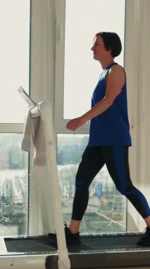 Güneşli bir günde koşu bandında büyük pencereye karşı yürüyen bir kadın. Koşu bandında spor yapan formda bir bayan. Evde fitness. Dikey video