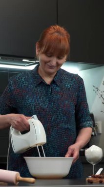 Mutfaktaki kadın elektrikli el mikseriyle beyaz krema çırpıyor. Dişi kek pişiriyor. Baker lezzetli bir pasta ya da pasta hazırlıyor. Ev yapımı fırın. Dikey video