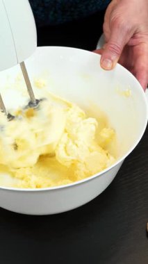 Kadın, modern mutfakta kremalı pasta yapıyor. Kadın fırıncı tereyağı ve şekeri elektrikli mikserle çırpıyor. Geleneksel ev yapımı pasta hazırlama süreci. Dikey video
