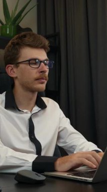 Adam ofiste dizüstü bilgisayarda çalışıyor, klavyede daktilo kullanıyor, bilgisayar faresi kullanıyor. İş yerinde çalışan gömlekli ve gözlüklü genç bir erkek. Dikey video