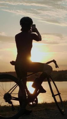 Gölün kenarında duran ve gün batımında akıllı telefondan fotoğraf çeken genç bir bisikletçinin silueti. Dişi sporcu gün batımında gölün yanında duruyor. Dikey video