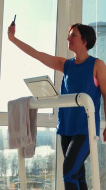 Çekici kadın evde koşu bandında yürürken akıllı telefondan selfie çekiyor. Genç, sıska kadın, evde akıllı eğitmen üzerinde egzersiz yapıyor. Dikey video