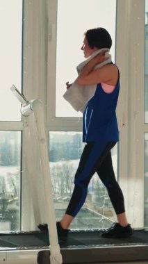 Evde koşu bandında koştuktan sonra yüzü havluyla silen formda bir kadın. Atletik güdümlü kadın koşan makinenin üzerinde yürüyor ve evde sağlıklı kalıyor. Dikey video